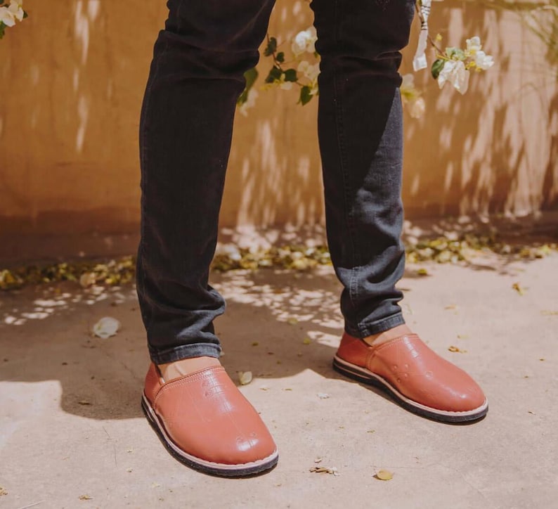 May include: A pair of slip-on shoes in a muted terracotta color with a smooth, rounded toe design and a thin, dark-colored sole. The shoes feature a low-cut opening and a simple, unadorned exterior surface, worn with dark-colored denim jeans.
