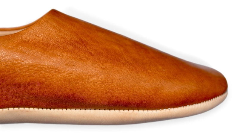 Peut inclure: Gros plan d'une pantoufle en cuir marron. La chaussure a un bout arrondi et une semelle de couleur claire. Le cuir a un aspect textur&eacute; et une couleur chaude et naturelle. La semelle est cousue &agrave; la tige.