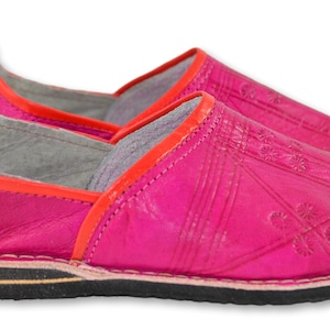 May include: A pair of bright pink leather slippers featuring a vibrant orange trim along the collar. The upper surface displays subtle embossed geometric patterns and floral motifs. The footwear includes a flat black rubber sole with a thin tan layer.