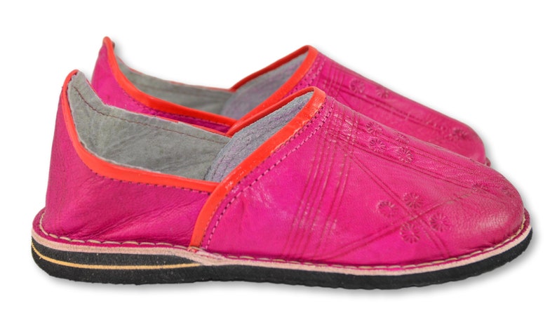 May include: A pair of bright pink leather slippers featuring a vibrant orange trim along the collar. The upper surface displays subtle embossed geometric patterns and floral motifs. The footwear includes a flat black rubber sole with a thin tan layer.