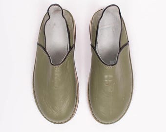 May include: A pair of olive green leather slippers. The slippers have a rounded toe, a low profile, and a black trim around the opening. The interior is white. The soles are a light brown color.