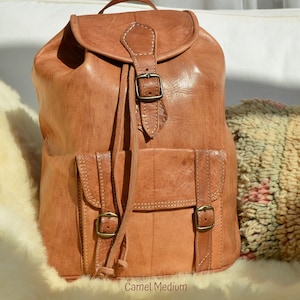 May include: A camel brown leather backpack with a flap top closure and two front pockets. The backpack has a buckle closure and adjustable straps.  The text "Camel Medium" is printed on the bottom of the image.