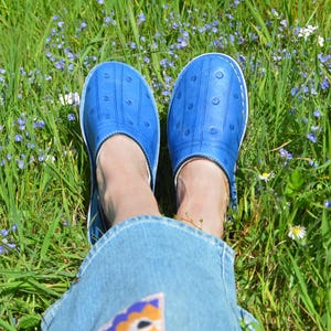 May include: A pair of bright blue slip-on clogs featuring a textured surface with small, evenly spaced circular indentations and vertical lines. The shoes have a white sole edge and white stitching detail around the perimeter of the upper section.