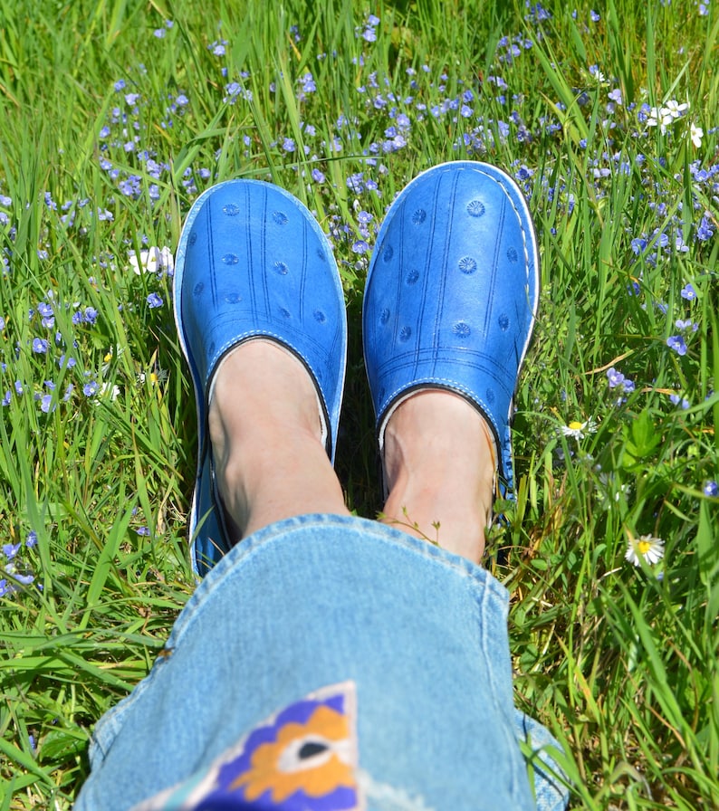 May include: A pair of bright blue slip-on clogs featuring a textured surface with small, evenly spaced circular indentations and vertical lines. The shoes have a white sole edge and white stitching detail around the perimeter of the upper section.
