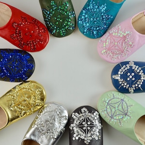Puede incluir: Un conjunto de diez zapatillas de cuero de colores con adornos de joyas en forma de copo de nieve. Las zapatillas est&aacute;n dispuestas en c&iacute;rculo sobre un fondo blanco.