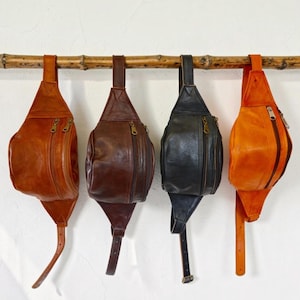 May include: Four leather bum bags hanging in a row, each featuring a single main compartment with a zippered closure and an adjustable strap. The bags are displayed in shades of tan, dark brown, black, and orange, all with metallic hardware.