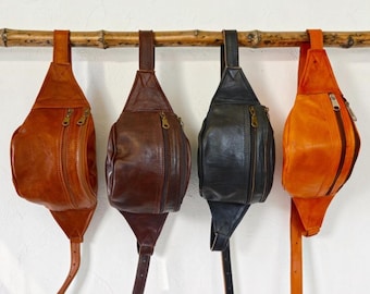 May include: Four leather bum bags hanging in a row, each featuring a single main compartment with a zippered closure and an adjustable strap. The bags are displayed in shades of tan, dark brown, black, and orange, all with metallic hardware.
