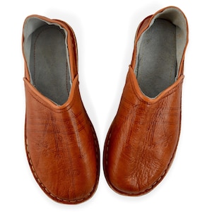 May include: A pair of tan leather slip-on shoes featuring a rounded toe, a low-cut opening, and a light gray interior lining. The exterior surface of the shoes displays a subtle embossed linear pattern with small floral details across the top.