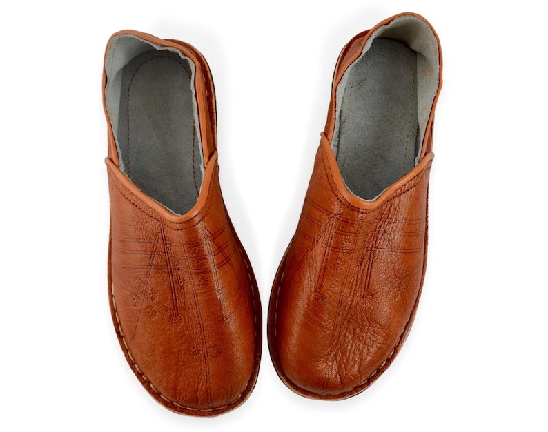 May include: A pair of tan leather slip-on shoes featuring a rounded toe, a low-cut opening, and a light gray interior lining. The exterior surface of the shoes displays a subtle embossed linear pattern with small floral details across the top.