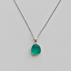 Pu&ograve; includere: Una collana d'argento con un piccolo ciondolo ovale color verde acqua. Il ciondolo &egrave; incastonato in una lunetta d'argento e appeso a una delicata catena d'argento. Lo sfondo &egrave; grigio chiaro.