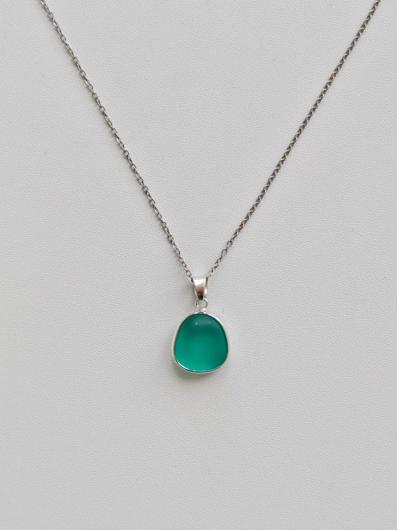 Pu&ograve; includere: Una collana d'argento con un piccolo ciondolo ovale color verde acqua. Il ciondolo &egrave; incastonato in una lunetta d'argento e appeso a una delicata catena d'argento. Lo sfondo &egrave; grigio chiaro.