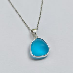 Pu&ograve; includere: Collana d'argento con un ciondolo. Il ciondolo ha una pietra blu chiaro, arrotondata e triangolare, incastonata in una lunetta d'argento. La pietra pende da una delicata catena d'argento. Il ciondolo ha una superficie liscia e lucida.