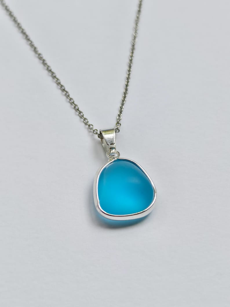 Pu&ograve; includere: Collana d'argento con un ciondolo. Il ciondolo ha una pietra blu chiaro, arrotondata e triangolare, incastonata in una lunetta d'argento. La pietra pende da una delicata catena d'argento. Il ciondolo ha una superficie liscia e lucida.