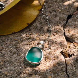 Pu&ograve; includere: Una collana d'argento con un ciondolo verde acqua. Il ciondolo ha una forma liscia e arrotondata con una lunetta d'argento. La delicata catena poggia su una superficie grigia strutturata. Una foglia gialla &egrave; sullo sfondo.