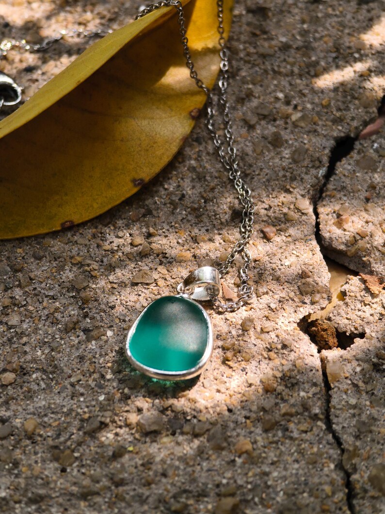 Pu&ograve; includere: Una collana d'argento con un ciondolo verde acqua. Il ciondolo ha una forma liscia e arrotondata con una lunetta d'argento. La delicata catena poggia su una superficie grigia strutturata. Una foglia gialla &egrave; sullo sfondo.