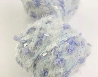 Pode incluir: Um novelo de fio macio e felpudo em tons de azul claro e branco com detalhes de glitter prateado. O fio tem uma textura felpuda e é perfeito para tricotar ou fazer crochê.