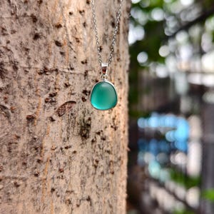 Pu&ograve; includere: Una collana d'argento con un pendente con una pietra arrotondata di colore verde acqua. La collana &egrave; appesa sullo sfondo di un tronco d'albero, con uno sfondo verde sfocato.