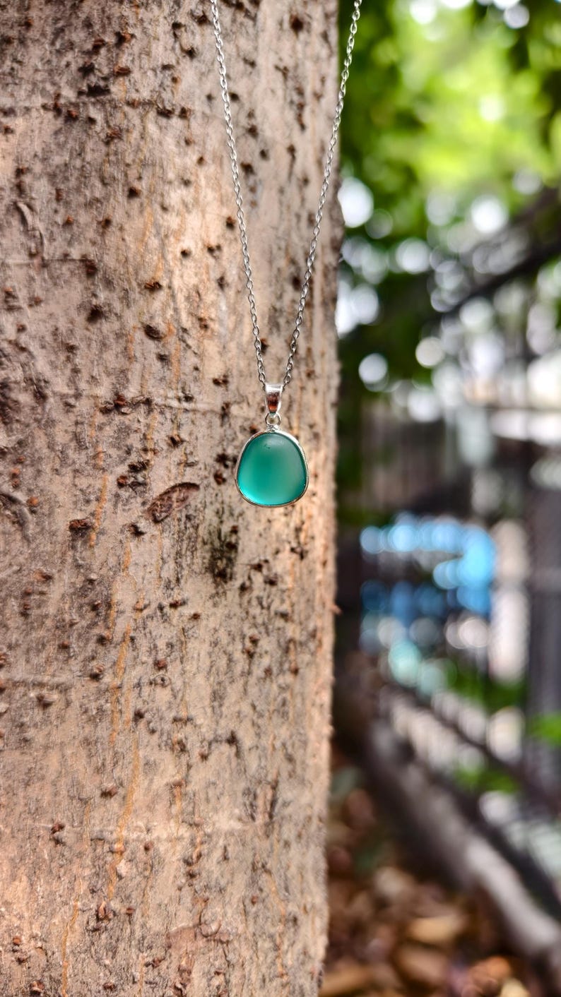 Pu&ograve; includere: Una collana d'argento con un pendente con una pietra arrotondata di colore verde acqua. La collana &egrave; appesa sullo sfondo di un tronco d'albero, con uno sfondo verde sfocato.