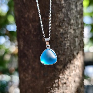 Pu&ograve; includere: Una collana d'argento con un ciondolo triangolare azzurro. Il ciondolo ha un aspetto smerigliato ed &egrave; incastonato in una lunetta d'argento. La catena &egrave; delicata, su uno sfondo naturale sfocato.