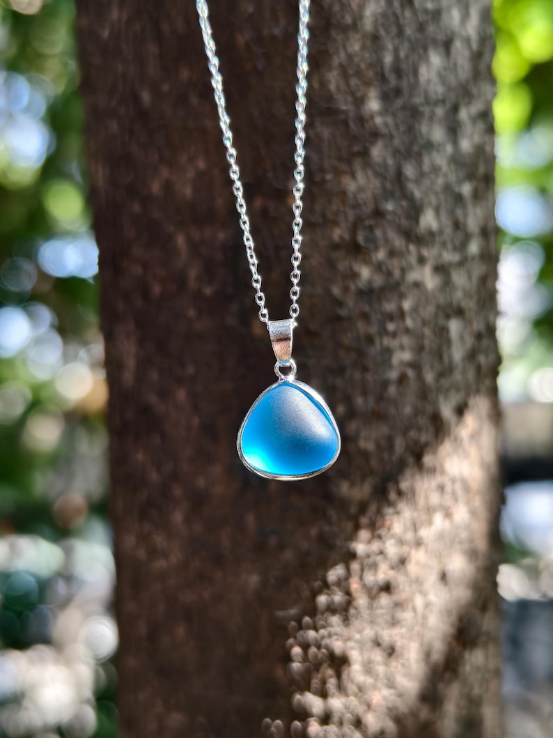 Pu&ograve; includere: Una collana d'argento con un ciondolo triangolare azzurro. Il ciondolo ha un aspetto smerigliato ed &egrave; incastonato in una lunetta d'argento. La catena &egrave; delicata, su uno sfondo naturale sfocato.