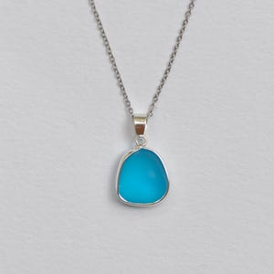 Pu&ograve; includere: Una collana d'argento con un ciondolo. Il ciondolo presenta una pietra turchese brillante e arrotondata, incastonata in una cornice d'argento. Pende da una delicata catena d'argento. Lo sfondo &egrave; di colore chiaro.