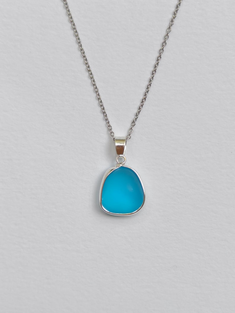 Pu&ograve; includere: Una collana d'argento con un ciondolo. Il ciondolo presenta una pietra turchese brillante e arrotondata, incastonata in una cornice d'argento. Pende da una delicata catena d'argento. Lo sfondo &egrave; di colore chiaro.