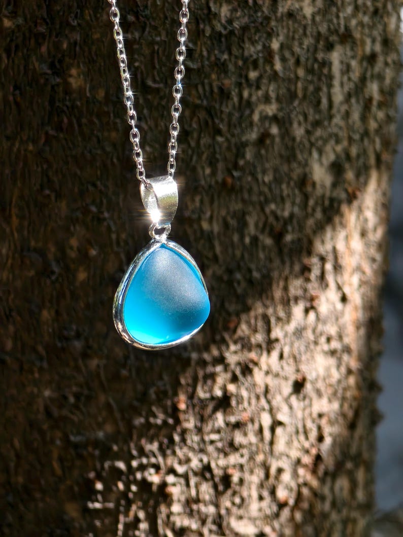 Pu&ograve; includere: Una collana d'argento con un ciondolo triangolare azzurro. Il ciondolo ha un aspetto liscio e smerigliato, incastonato in una lunetta d'argento. La catena &egrave; delicata, appesa su uno sfondo sfocato e strutturato.