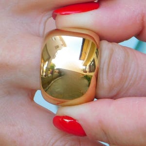 May include: A wide, polished gold-tone band ring with a smooth, reflective surface. The metal surface acts as a mirror, displaying a clear reflection of a building exterior and outdoor surroundings on its curved, high-shine exterior finish.