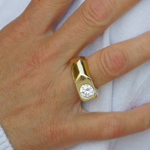 May include: A gold ring with a large, clear, round gemstone set in the center. The ring has a thick band with a geometric design.