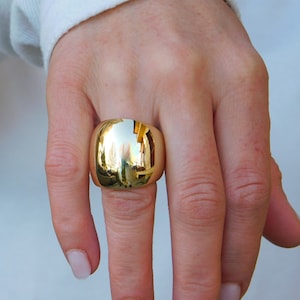 May include: A large, gold-colored ring with a rounded, dome-shaped design. The ring is worn on the index finger of a hand.