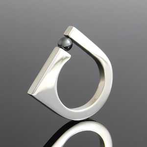 May include: A modern silver ring with a unique teardrop design. The ring features a polished finish and a small, dark gray sphere set at the top. The ring's smooth, curved lines and reflective surface create a minimalist aesthetic.