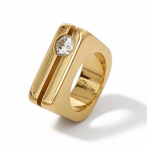 May include: A polished gold ring featuring a thick, rectangular band with a vertical channel running down the center. A single, round, clear faceted gemstone is set securely within the top portion of the channel, catching the light.