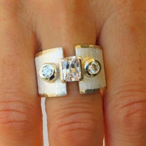 Puede incluir: Un anillo ancho de tono plateado con acabado de metal cepillado, que presenta una piedra preciosa rectangular transparente central flanqueada por dos piedras redondas más pequeñas, engastadas en biseles lisos sobre la banda.