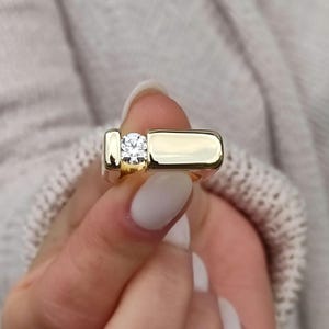 May include: A gold band ring with a single, round, clear diamond set in the center.