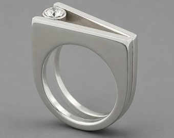 Può includere: Un anello in metallo color argento caratterizzato da una fascia larga con parte superiore piatta e un'apertura a forma di V sulla corona. Una singola gemma rotonda e trasparente è incastonata nell'angolo interno dell'apertura a V.