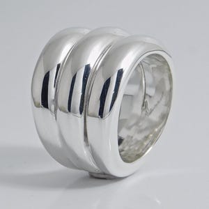 May include: A wide, polished silver ring featuring a triple-band design. The ring is composed of three smooth, rounded, parallel silver segments that are joined together to create a single, substantial, and reflective metallic band.