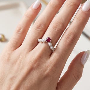 May include: A silver ring with a central, round, red gemstone, surrounded by small white pearls. The ring is on a hand with manicured nails, set against a white background with other jewelry.