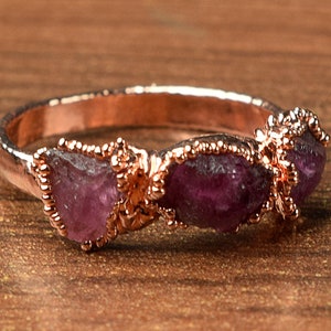 May include: A rose gold ring with three raw amethyst stones set in the band. The stones are a deep purple color and have a natural, rough texture.