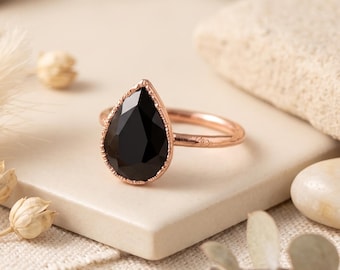 May include: A rose gold ring featuring a faceted, pear-shaped black gemstone set in a delicate, beaded metal frame. The band is smooth and circular, showcasing a polished finish that complements the dark, reflective surface of the central stone.