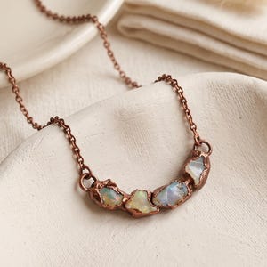 May include: A necklace featuring a copper-toned chain and a curved pendant composed of four raw, iridescent opal stones set in textured copper metal. The stones display flashes of blue, green, and white light against the metallic setting.