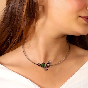 May include: A delicate necklace featuring a thin, bronze-colored band. The pendant showcases a cluster of gemstones in shades of green and pink. The necklace is worn by a person wearing a white top.