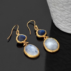 Può includere: Un paio di orecchini pendenti in oro con due pietre ciascuno. La pietra superiore è un ovale blu scuro, e la pietra inferiore è un ovale bianco con una finitura scintillante.