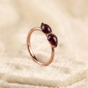 Könnte beinhalten: Ein Ring aus Roségold mit zwei tropfenförmigen, tiefroten Granat-Edelsteinen, die nebeneinander angeordnet sind. Der Metallring ist glatt und poliert, während die Fassungen der beiden dunkelroten Steine eine feine, perlenartige Textur aufweisen.
