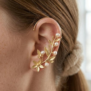 Puede incluir: Puño de oreja dorado con un diseño floral y de hojas, adornado con pequeñas perlas blancas redondas. El puño se curva alrededor de la oreja, con una flor en la base y una delicada estructura en forma de vid que se extiende hacia arriba.