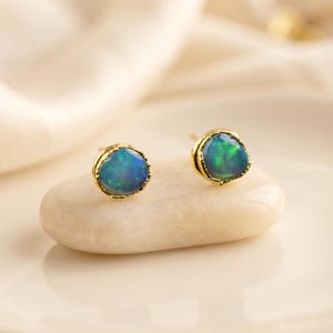 May include: A pair of stud earrings featuring round, iridescent blue and green opals set in textured gold metal bezels. The stones display a play of color with flashes of teal and emerald, secured by gold posts, showcasing a polished finish.