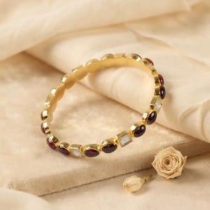 Peut inclure: Un bracelet jonc doré orné d'un motif alterné de pierres cabochons rouge foncé rondes et de petites pierres claires de taille carrée, serties tout autour de la bande métallique pour créer un design répétitif et symétrique.