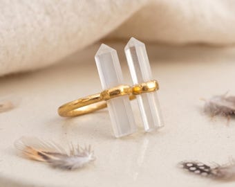 Könnte beinhalten: Ein goldfarbener Ring mit zwei vertikalen, klaren, hexagonal geschliffenen Kristallspitzen, die nebeneinander auf einem dünnen, polierten Metallband montiert sind. Das Design zeigt eine symmetrische Anordnung der beiden durchscheinenden Steinsäulen.