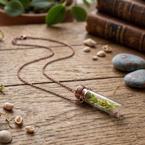 Peut inclure: Un collier composé d'un petit pendentif en forme de fiole en verre transparent, surmonté d'un bouchon en métal cuivré. À l'intérieur de la fiole, un petit brin de plante verte est visible. Le pendentif est suspendu à une chaîne en métal cuivré.