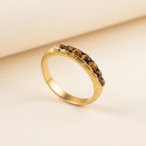 Può includere: Un anello color oro con una fila di piccole pietre preziose occhio di tigre, rotonde e sfaccettate, incastonate lungo la metà superiore della fascia. L'anello in metallo ha una finitura opaca liscia e una texture organica irregolare.