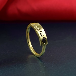 May include: A gold-coloured ring with the name "ELENA" engraved on it. The ring has a small heart shape at the end of the engraved name.